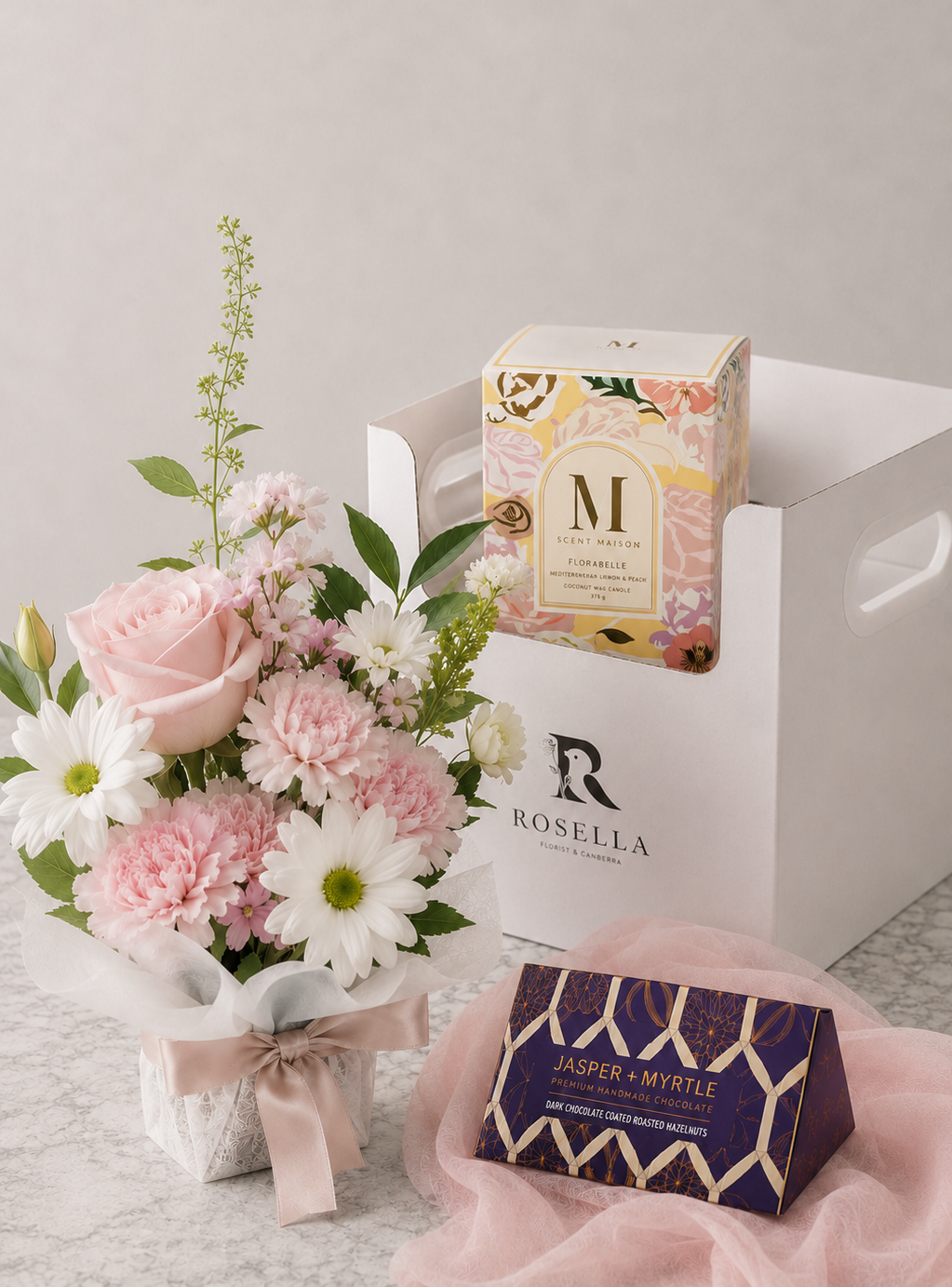 Fresh flower arrangement paired with a scented candle displayed on a white gift box and a box of dark chocolate, creating a premium gift set from Rosella Florist Canberra.