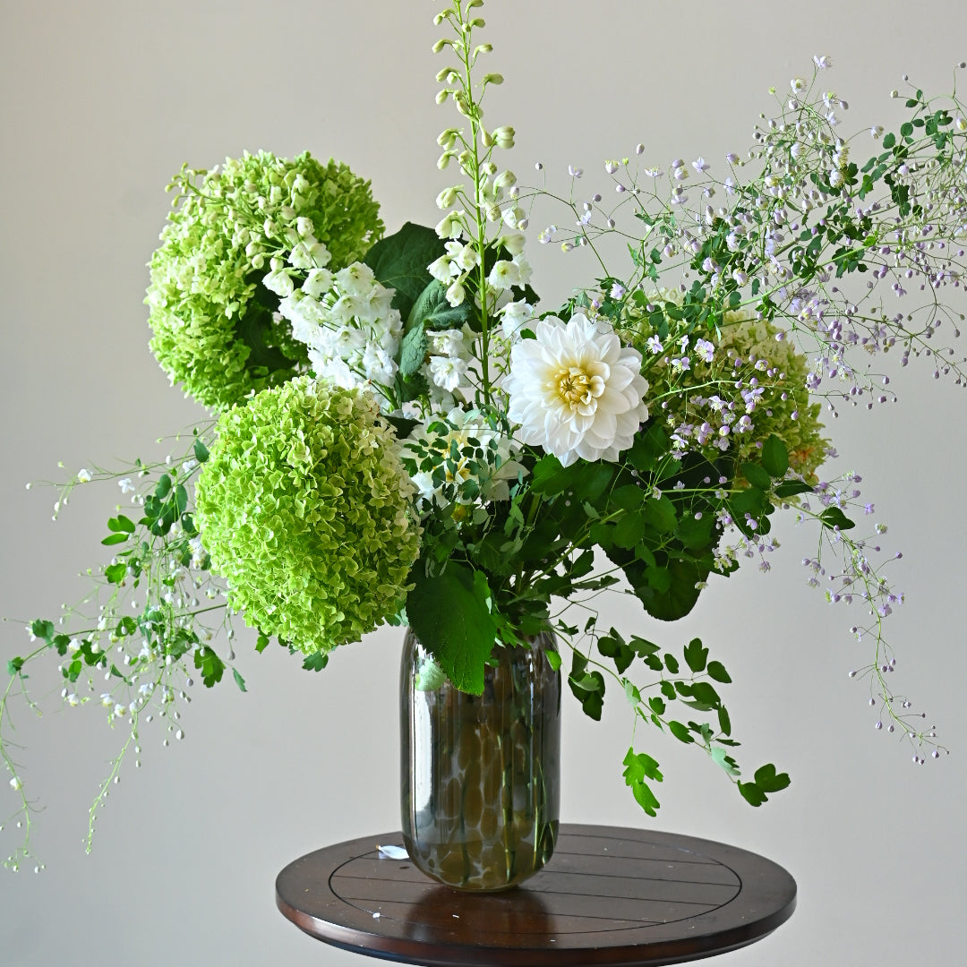 A floral arrangement featuring green and white flowers, including what appears to be round and long-stemmed flowers, arranged in a glass vase placed on a wooden surface against a neutral background.