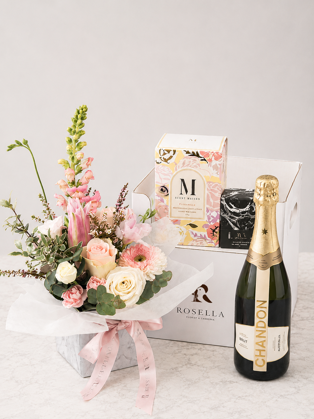 Fresh flower arrangement with a bottle of sparkling wine and two scented candles displayed on a white Rosella gift box, creating a premium gift hamper from Rosella Florist Canberra.
