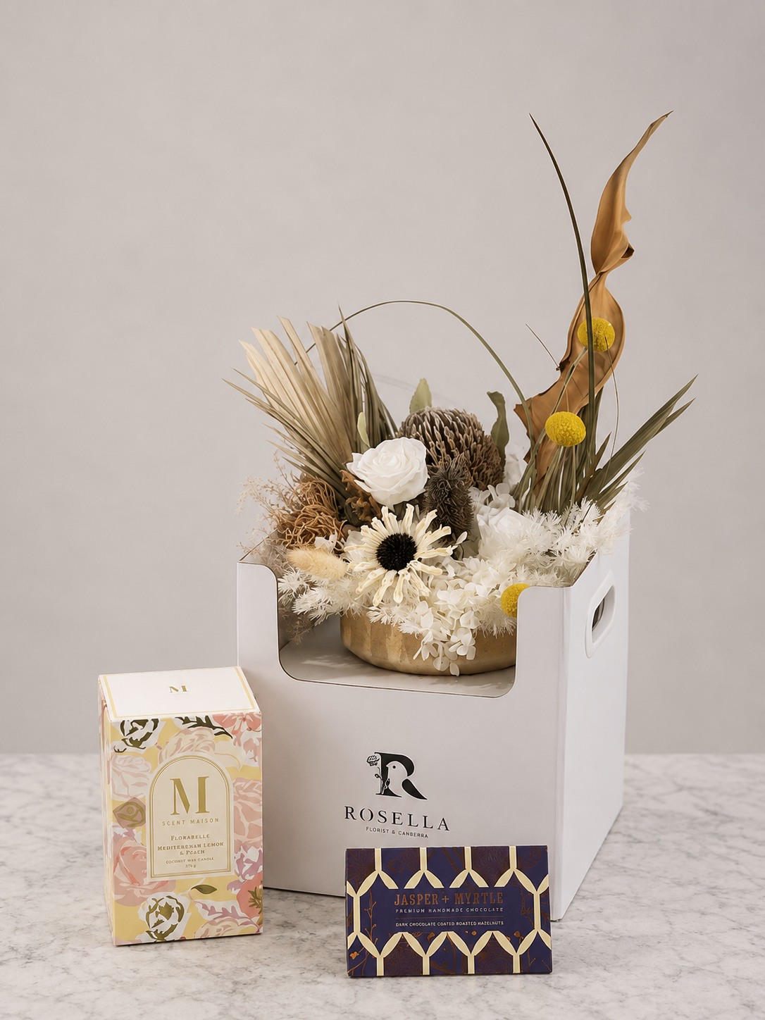 Gift set featuring a scented candle, preserved flower arrangement displayed on a white gift box and two boxes of artisan chocolate from Rosella Florist Canberra.