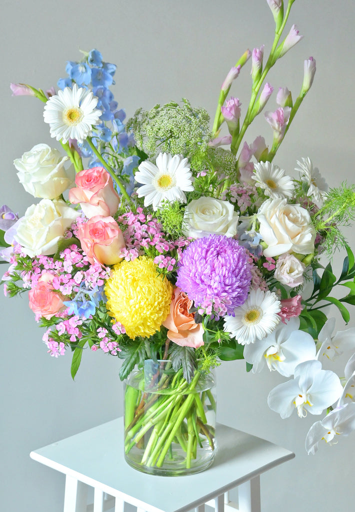 Large pastel flower arrangement in a glass vase featuring pink and white roses, white orchids, purple and yellow chrysanthemums, light blue delphiniums, pink gladiolus and seasonal blooms, handcrafted by Rosella Florist Canberra.